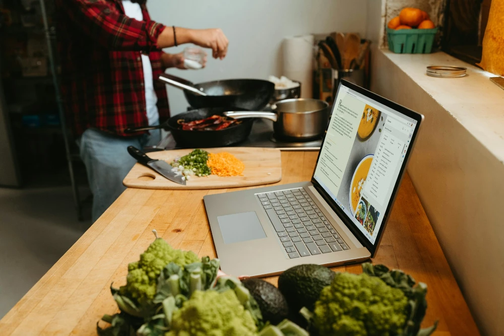 Photo of a man cooking with an open laptop on the counter, with a recipe on the screen.